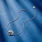 A delicate silver chain bracelet featuring a circular tree of life pendant with small clear stones, displayed on a textured blue fabric background.