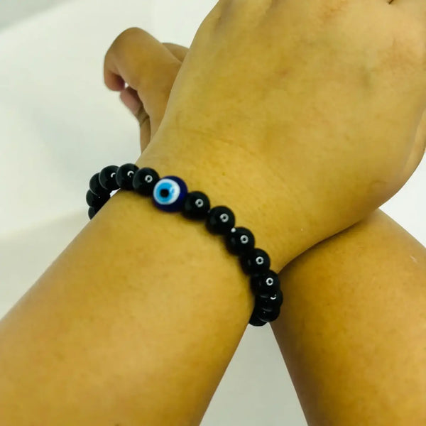 A close-up of a wrist wearing a black beaded bracelet featuring small black beads with white accents. At the center is a larger bead with a blue and white evil eye design, symbolizing protection.