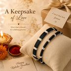 Two black braided Raksha Bandhan bracelets with silver metallic accents displayed on a beige cushion, alongside a gift box, lit candle, flowers, and greeting cards with the text 'A Keepsake of Love' and 'Happy Raksha Bandhan'.