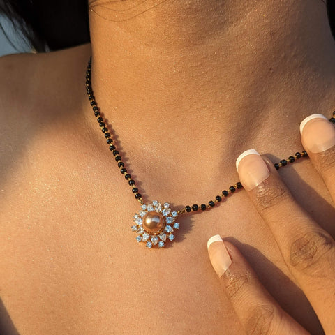 A traditional yet contemporary mangalsutra design featuring a black beaded chain made of 92.5 sterling silver with gold plating, a genuine freshwater pearl pendant surrounded by cubic zirconia stones as the centerpiece, worn on a woman's hand against a skin-toned background.