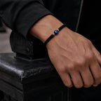 A close-up of a person's hand resting on a black metal surface, wearing a black braided cord bracelet with a blue evil eye charm.