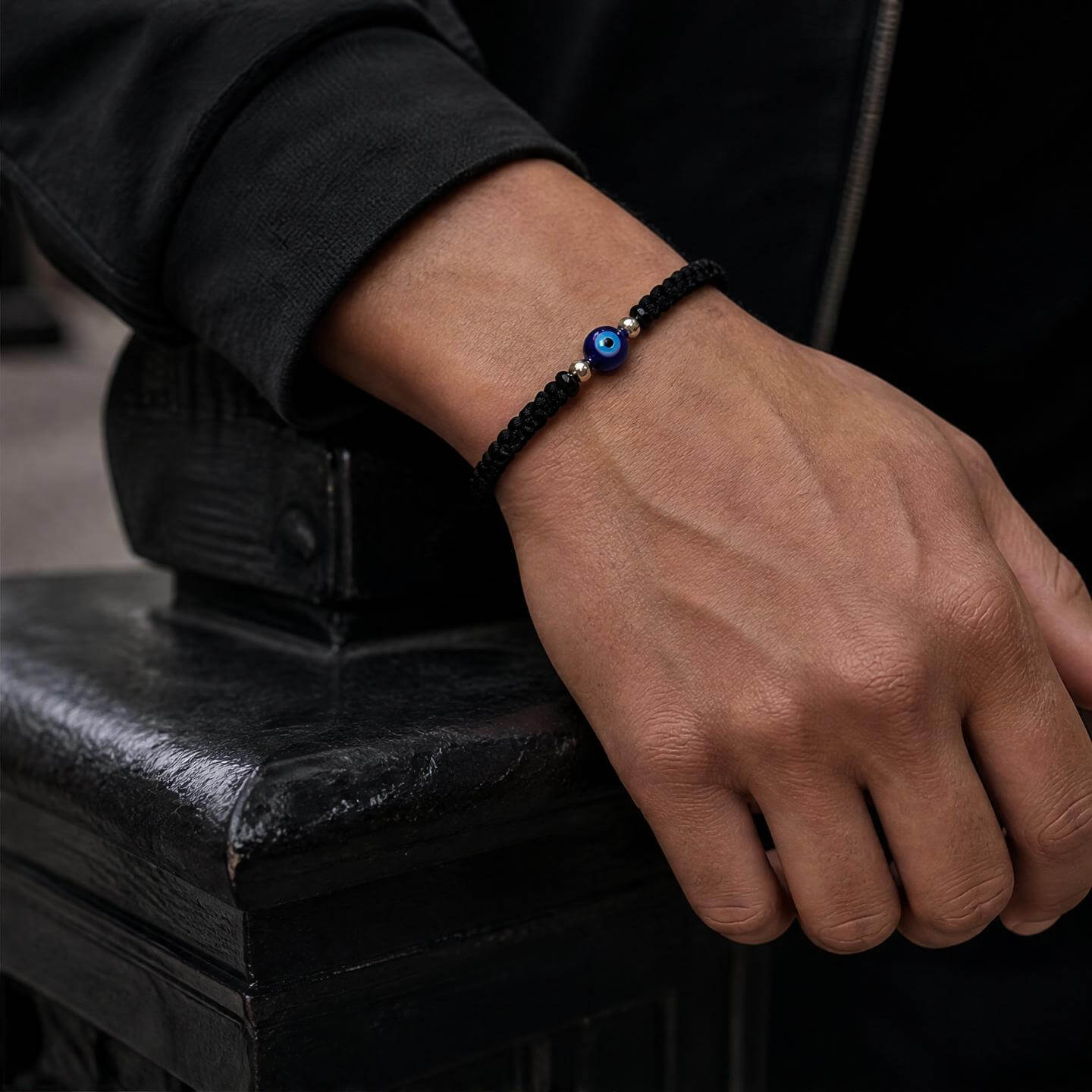 A close-up of a person's hand resting on a black metal surface, wearing a black braided cord bracelet with a blue evil eye charm.