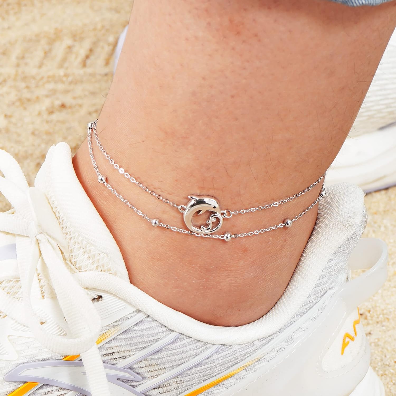 Silver anklet with a dolphin charm on a person's ankle against a sandy background